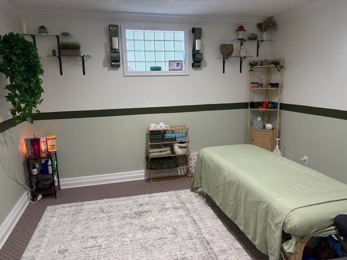A spa treatment room with a massage table covered in cream fabric, light wood shelving, potted plants, wall-mounted speakers, and a window with a black accent stripe on cream walls.